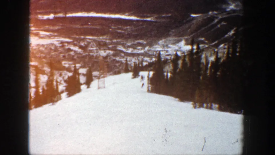 1961 Aspen home-movie frame with a skier on a broad snow-covered run, evergreen trees to the side, and a distant valley under a bright sky.