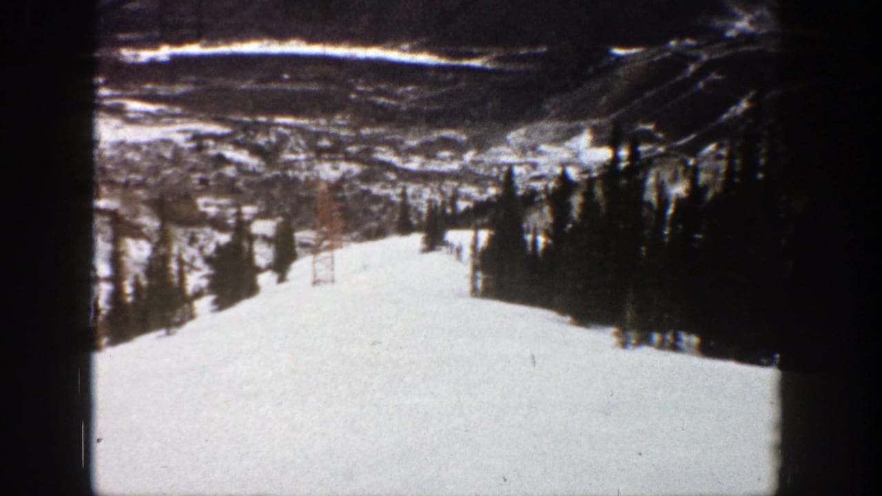 Snowy ski slope with evergreen trees, mountain ridges, and lift structures in the distance.