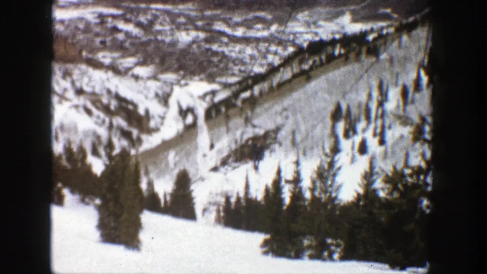 1961 home-movie frame of Aspen ski terrain with snow-covered slopes, dark evergreens, and cable lift lines across the mountain.
