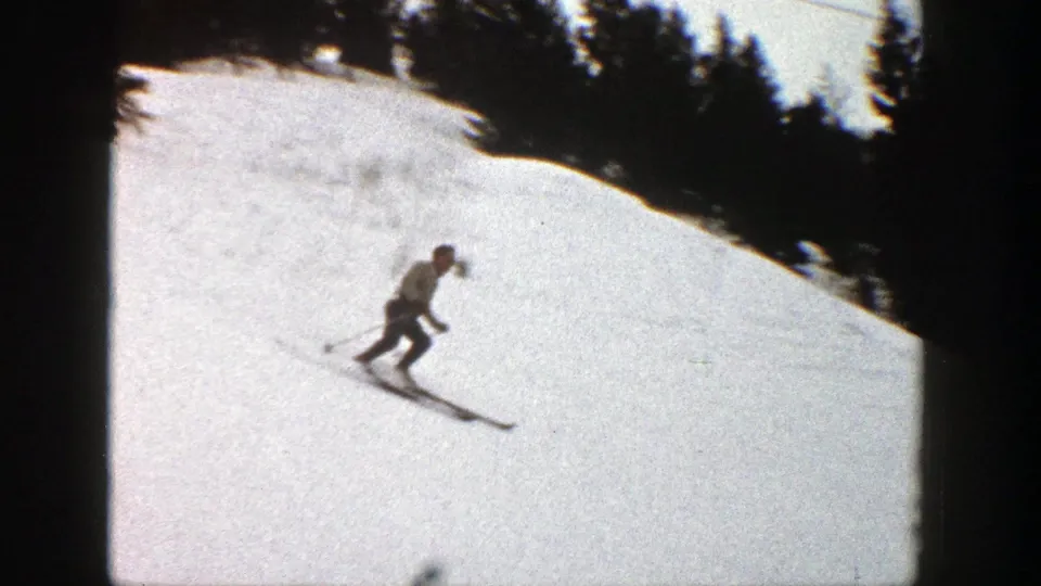 A skier in a light top and dark pants traverses the bright Aspen snowfield, with evergreen trees behind the slope.
