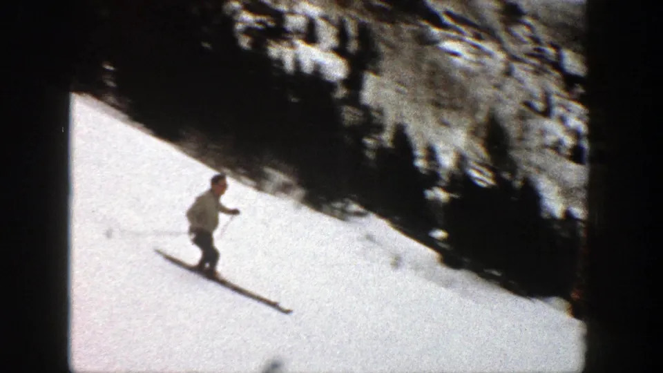 The skier continues downhill on the snow, passing near a dark shaded edge of trees or rock in the 1961 Aspen footage.
