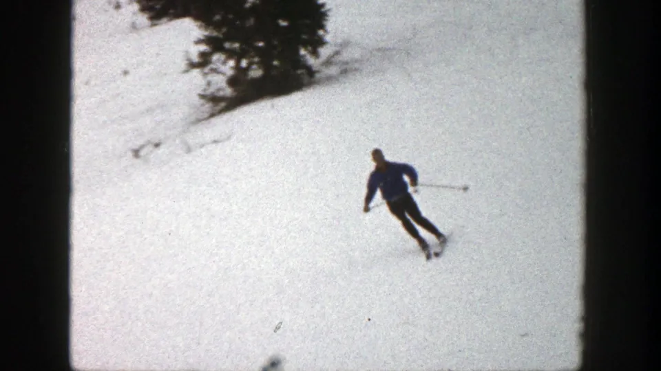 A skier in a blue jacket carves across the Aspen slope with dark trees visible above the snow.