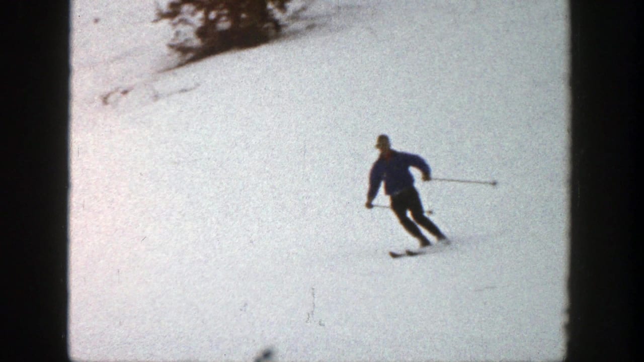 1961 Aspen home-movie frame showing a skier in a dark blue jacket carving across bright snow with poles extended.