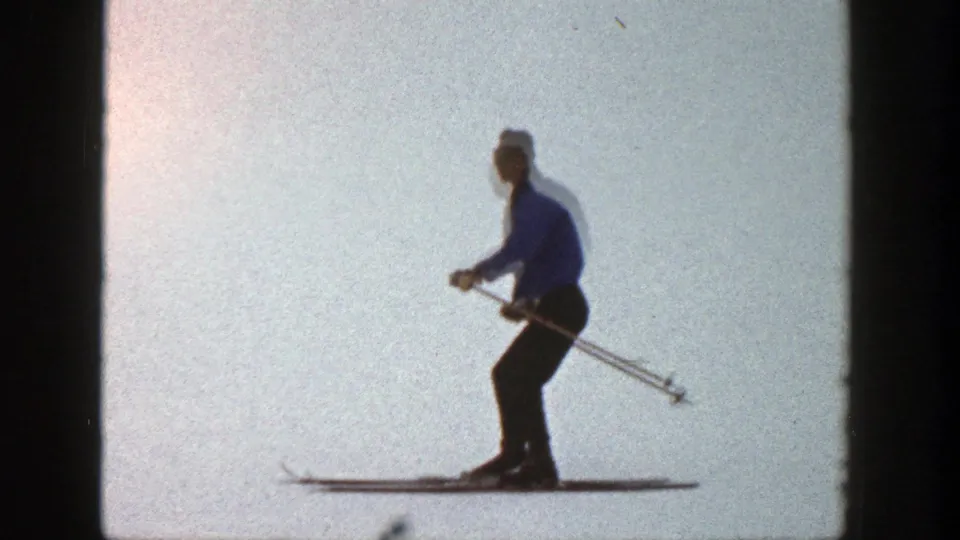 1961 Aspen home-movie frame with the skier in profile, poles angled back, crossing a bright snowy field.