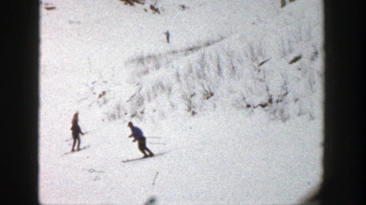 1961 Aspen home-movie frame showing two distant skiers on a sunlit snowy slope with exposed dark terrain above.