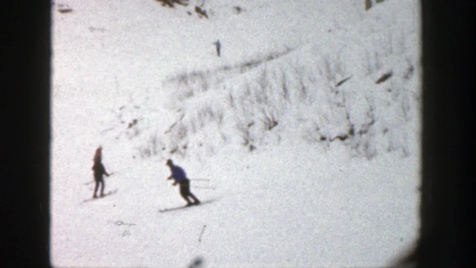 1961 Aspen home-movie frame showing two distant skiers on a sunlit snowy slope with exposed dark terrain above.