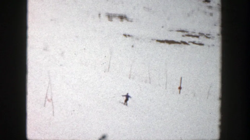 1961 Aspen home-movie frame showing a distant skier crossing a bright snowfield with visible 8mm grain and gate edges.