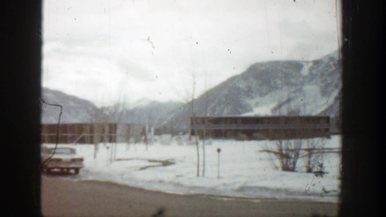 Snowy resort grounds with a parked car, low building, bare trees, and mountains behind.