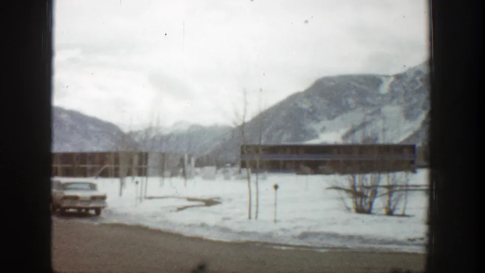 1961 Aspen winter frame with a classic car beside snowy lodge grounds and mountain slopes in the distance.