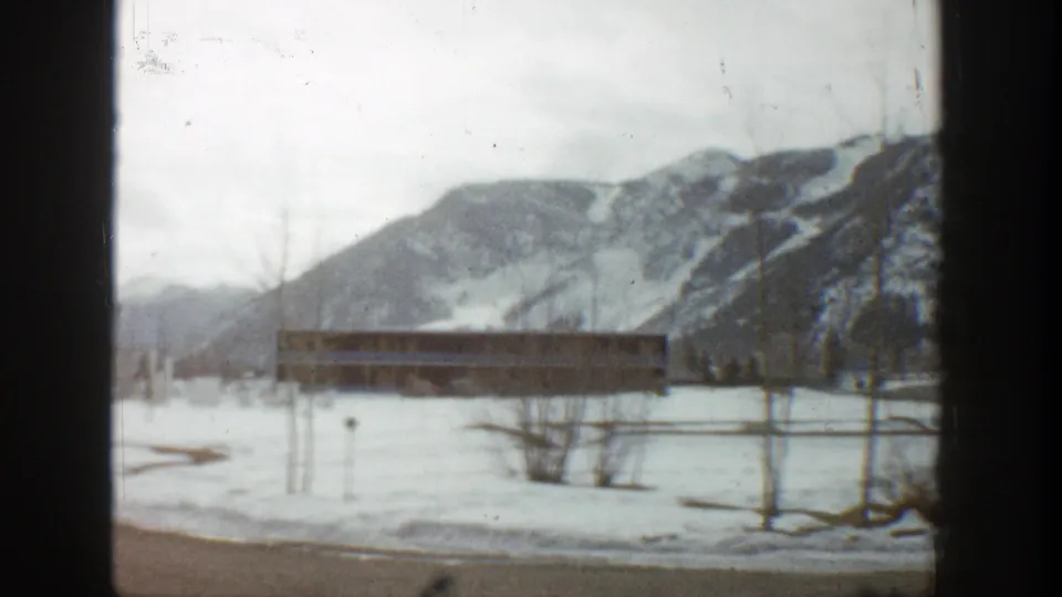 1961 Aspen, Colorado home-movie frame showing snow-covered ground, a long low building, bare trees, and distant mountains.