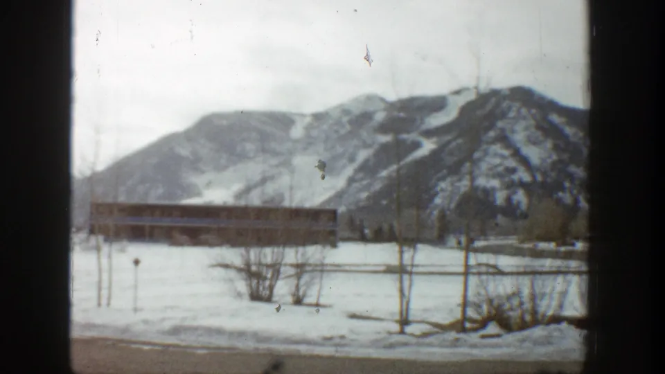 Aspen winter frame with a low structure across a snowy field, sparse bare trees, and snow-streaked mountains behind.