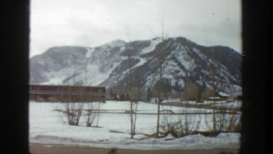 1961 Aspen home-movie view panning past a partial low building, roadside brush, snowfields, and dark mountain ridges.