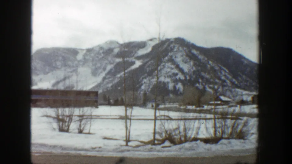 Late-frame 1961 Aspen winter view with bare shrubs and posts in front of snow-covered terrain and mountain slopes.