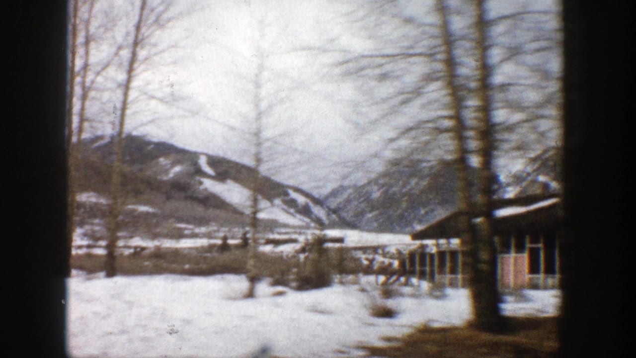 1961 Aspen-area home-movie frame showing snow, bare trees, low lodge buildings, and mountain slopes in the distance.