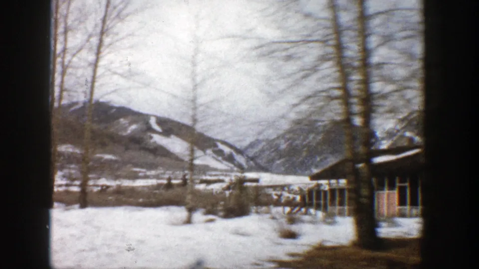 1961 Aspen-area home-movie frame showing snow, bare trees, low lodge buildings, and mountain slopes in the distance.