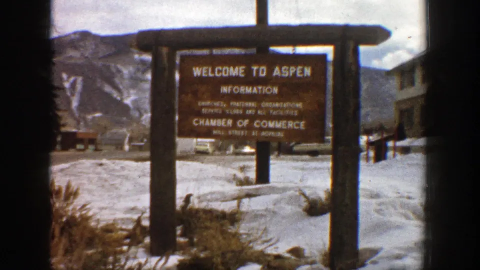 1961 Aspen home-movie frame centered on a wooden Welcome to Aspen information sign with snowbanks and mountains behind it.