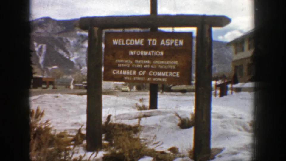 1961 Aspen home-movie frame of the wooden information sign framed by snow, parked cars, a building, and mountain slopes.
