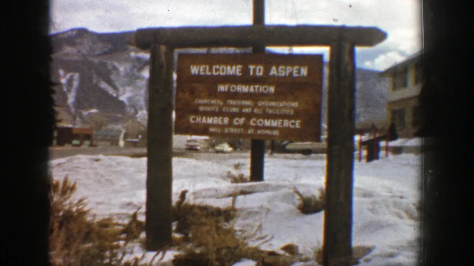 The Aspen welcome and information sign appears in a snowy 1961 mountain-town view with distant structures behind it.