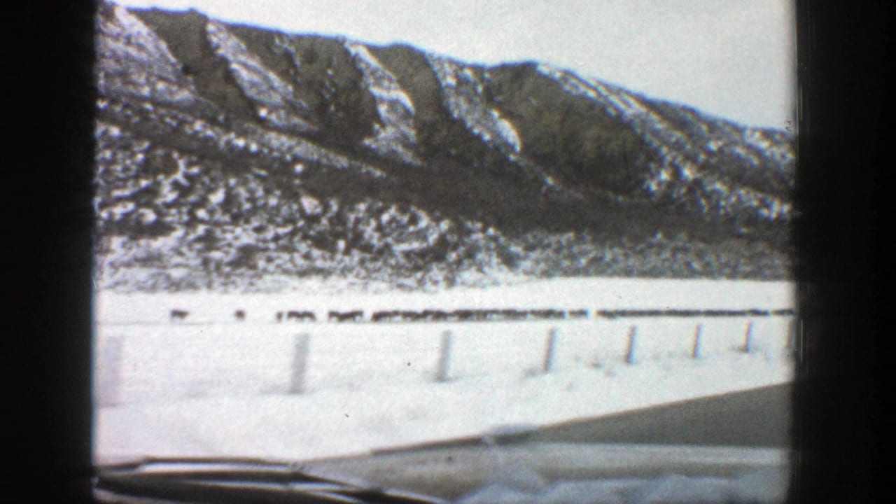 Snowy mountain road seen through a car windshield with guardrail posts and dashboard visible.