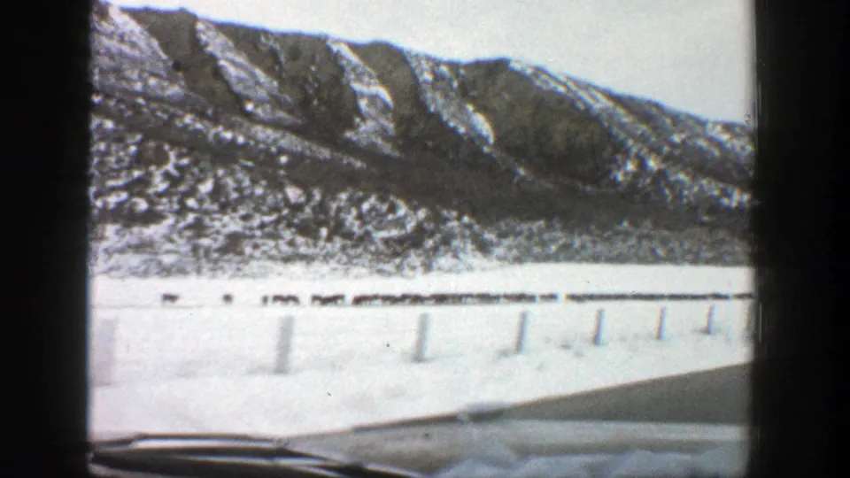 1961 Aspen, Colorado home-movie frame showing snow-covered mountains from inside a moving car, with windshield wipers and roadside posts visible.