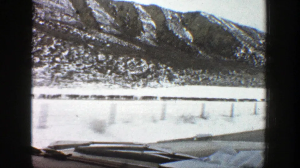 1961 Aspen, Colorado road-trip view with snow-dusted mountains, a flat snowy roadside, guardrail posts, and the car hood at the bottom of frame.