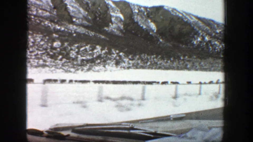 1961 Aspen, Colorado home-movie frame with the camera looking past the car hood toward snowy roadside posts and mountain slopes.
