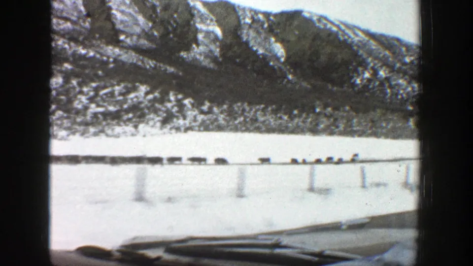 Late-frame 1961 Aspen, Colorado windshield view showing a snowy mountain slope, roadside guardrail posts, and the car hood in soft 8mm color.