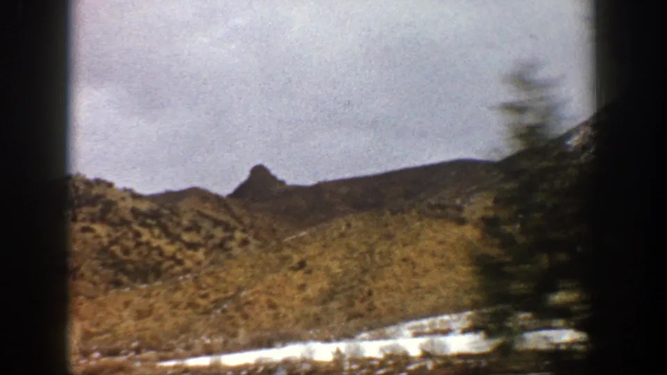 1961 8mm mountain view near Aspen with snowy ground, low hills, and a tall evergreen blurred by vehicle movement.