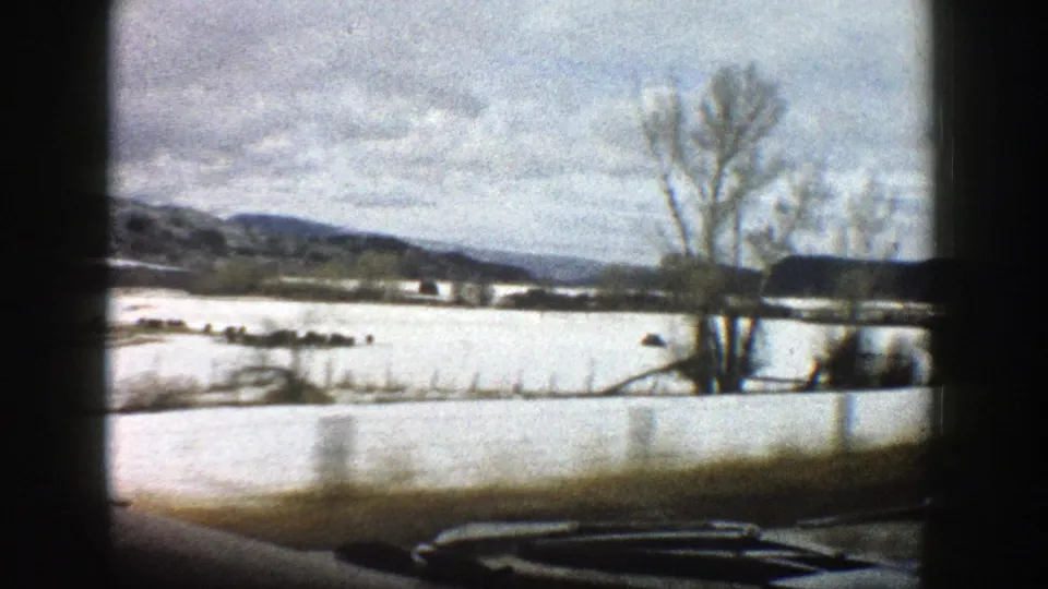 A moving-car view shows bare winter trees, snow-covered land, and low Colorado hills in soft 8mm color.