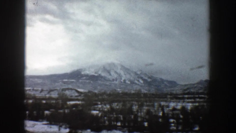 A cloudy 1961 Aspen-area mountain view shows snow-streaked slopes and dark winter trees from a traveling vehicle.