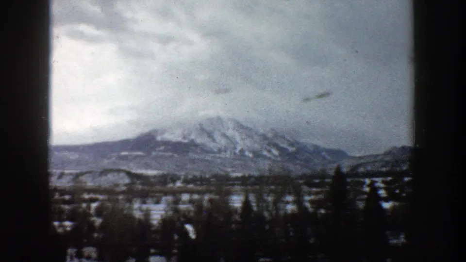 1961 Aspen, Colorado 8mm frame with a snow-dusted mountain range behind dark trees and open winter ground.