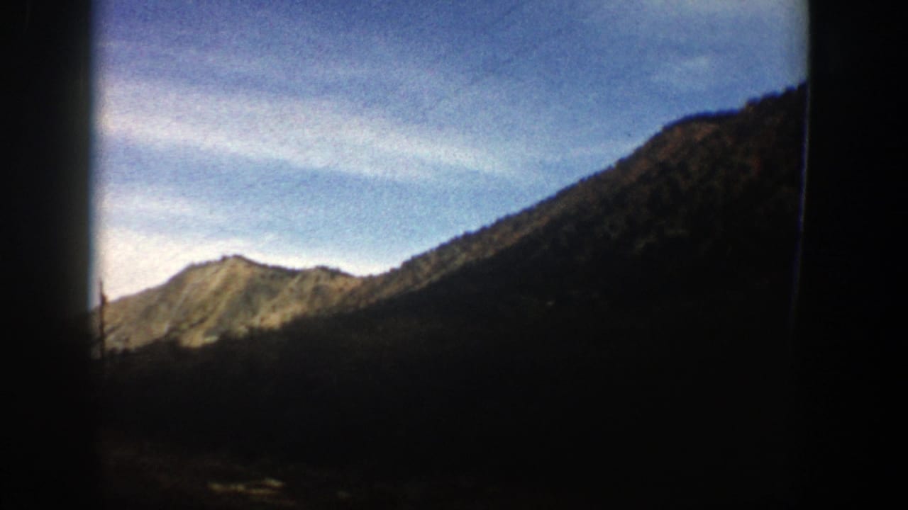 1961 Aspen, Colorado home-movie frame showing a mountain ridge, wooded slope, bright sky, and heavy shadow from a moving-view perspective.