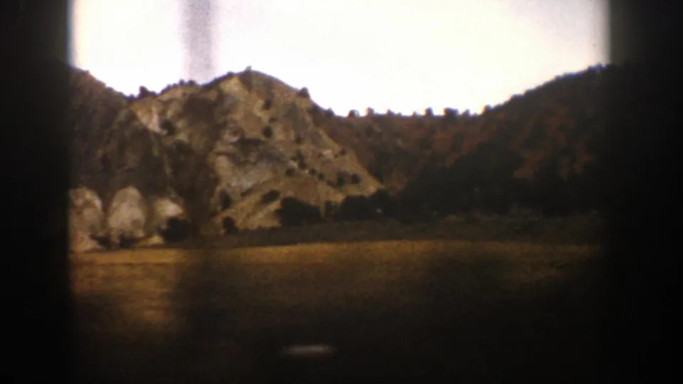 1961 Aspen, Colorado footage frame with a rocky hillside, open foreground, distant trees, and dark edge artifacts from the moving 8mm view.