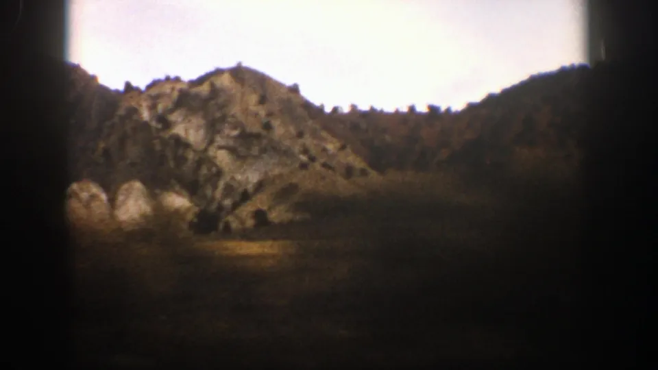 1961 Aspen, Colorado home-movie frame showing a rocky slope, large boulders, dark foreground shadow, and surrounding mountain terrain.