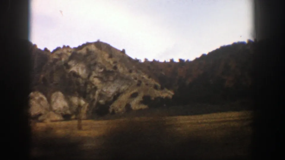 1961 Aspen, Colorado frame showing rocky hillside terrain, a low open foreground, distant trees, and dark film-edge shadows from the moving shot.