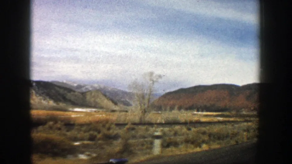 1961 Aspen, Colorado road footage frame with a bare tree, sagebrush flats, low hills, and mountains seen through the vehicle window.