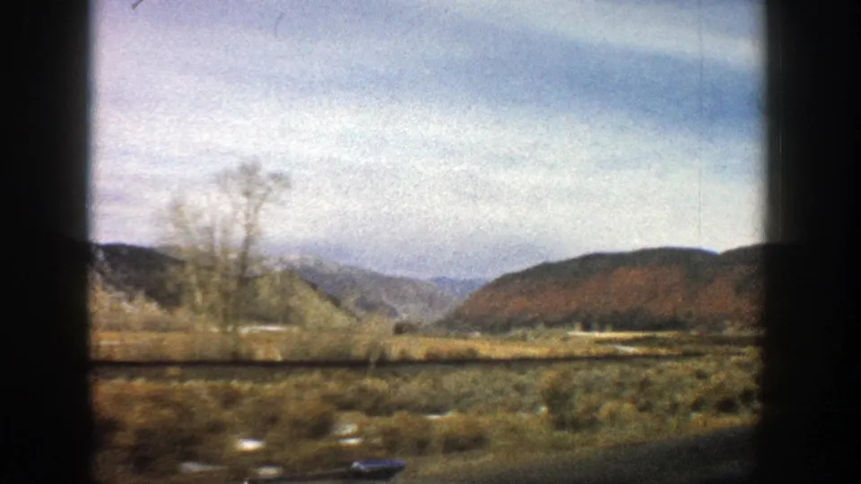 1961 Aspen, Colorado frame from a moving car, showing bare roadside trees, sagebrush terrain, low hills, and distant mountains.