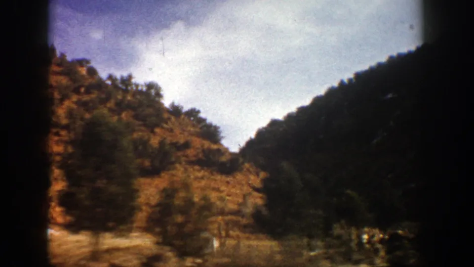 1961 Aspen-area 8mm view of a rocky hillside, trees, and bright sky from a vehicle on a mountain road.