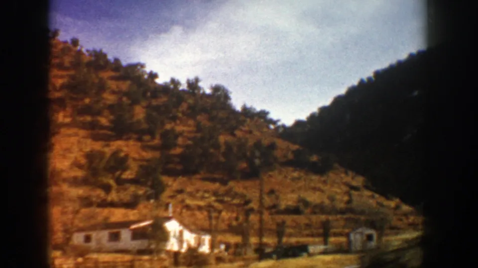 Aspen-area mountain view with a small white structure at the base of a rocky hillside, filmed from a moving vehicle in 1961.