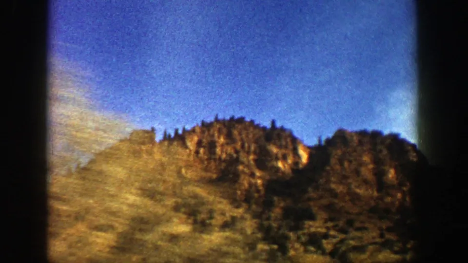 1961 8mm mountain landscape near Aspen, with a sunlit rocky ridge, grassy slope, trees, and blue sky.