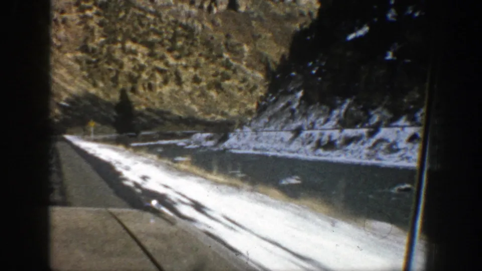 Aspen-area vehicle view of a snow-lined road beside water and steep rocky slopes, captured on 8mm film in 1961.