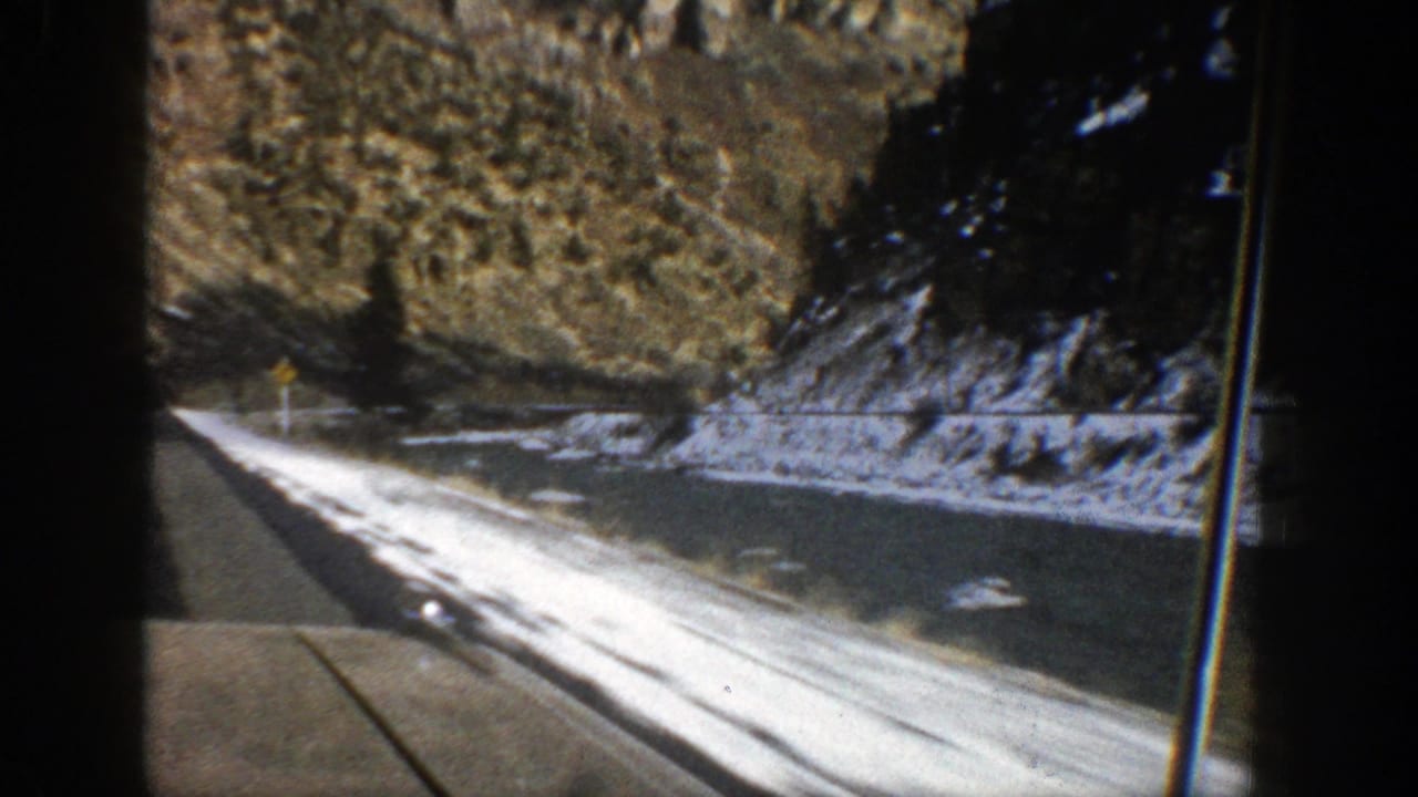 1961 Aspen-area home-movie view through a vehicle window of a snowy highway, icy shoulder, evergreen trees, and rocky mountain slope.