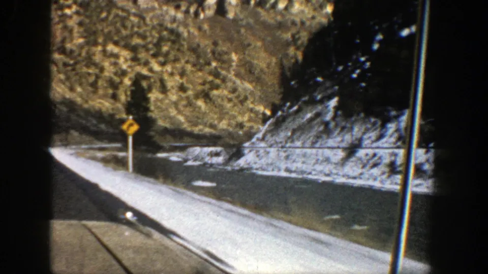 1961 Aspen, Colorado road scene with a yellow curve warning sign, snow along the pavement, evergreen trees, and a rocky slope.