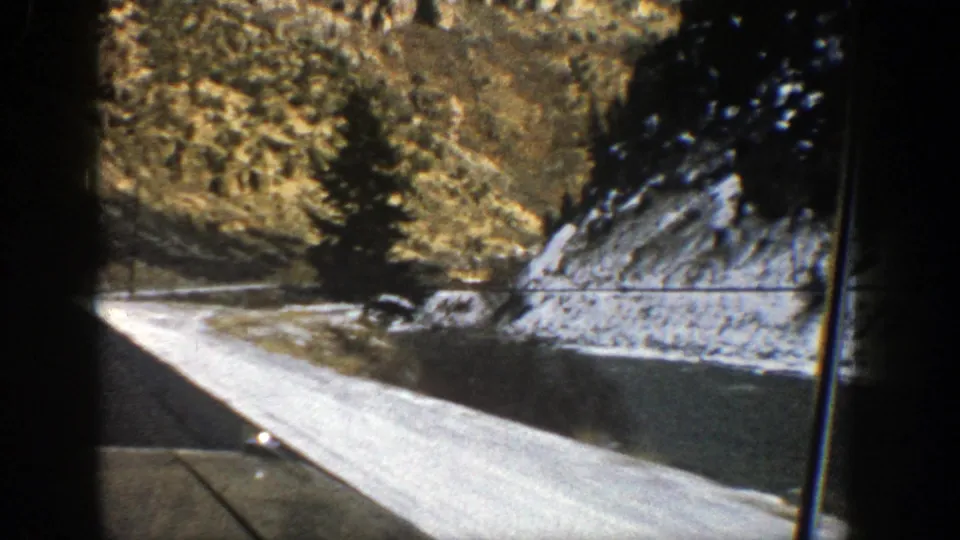 Passenger-view 8mm frame from 1961 Aspen showing a snow-covered roadside, dark pavement, evergreens, and a rocky mountain slope.