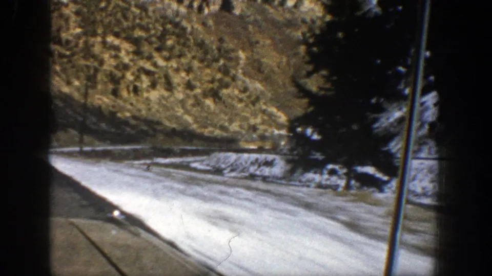 1961 Aspen-area home-movie frame of a winter highway curve, snowbanks, evergreen trees, and mountain terrain seen from a moving car.
