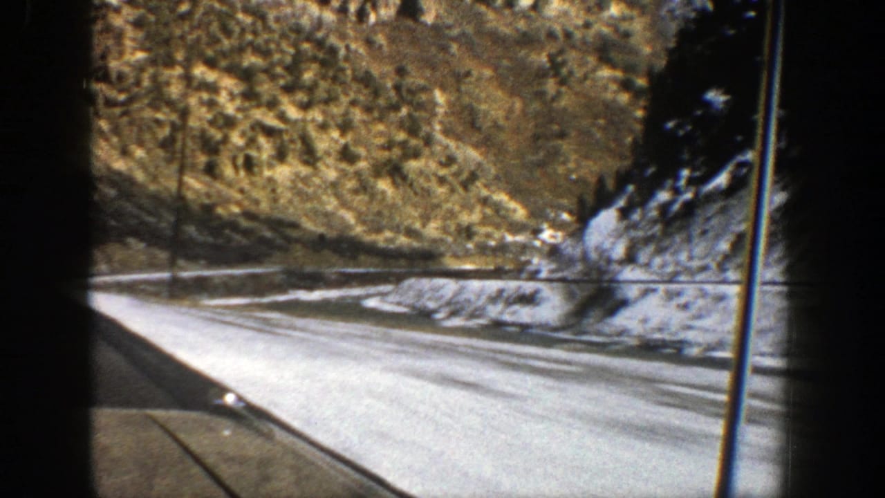 Passenger-window view of a mountain road beside snowy ground and rocky slopes.