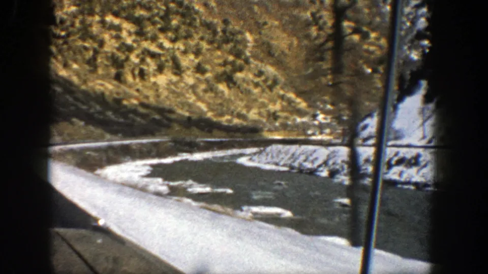 1961 Aspen, Colorado frame with a passenger-side view of snow patches, roadside pavement, and rugged mountain terrain beyond the window.