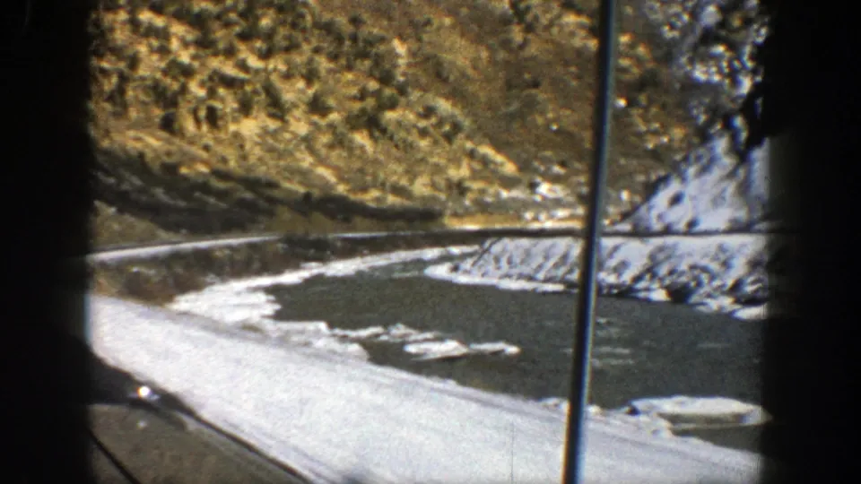 Final 1961 Aspen-area frame showing snow along the road, a dark ravine or watercourse, rocky slopes, and window framing from inside the car.