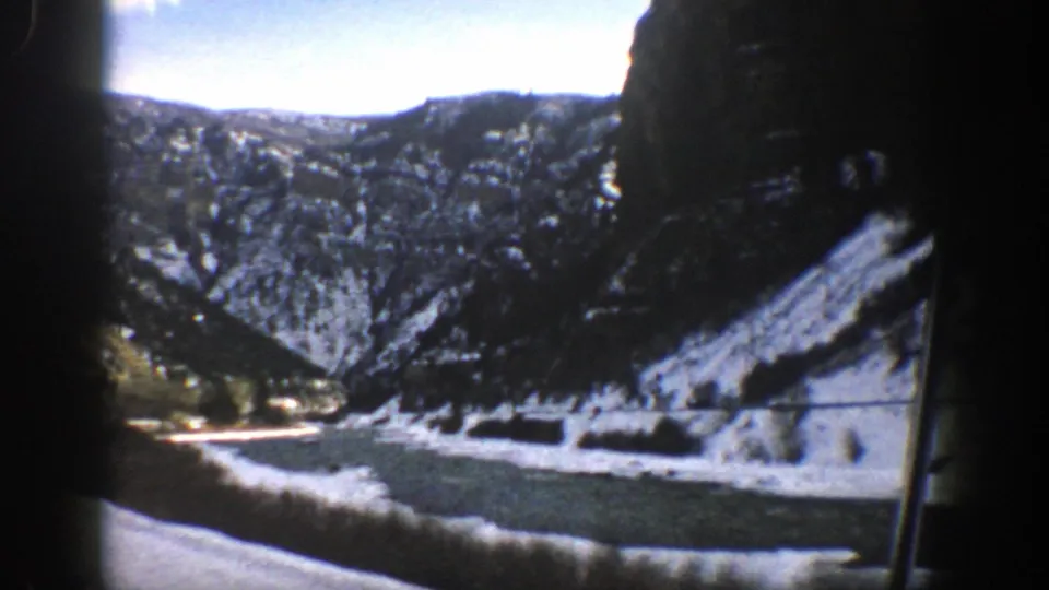 The Aspen-area road passes between snow-dusted mountains, with a waterway beside the route and dark interior or gate edges framing the view.