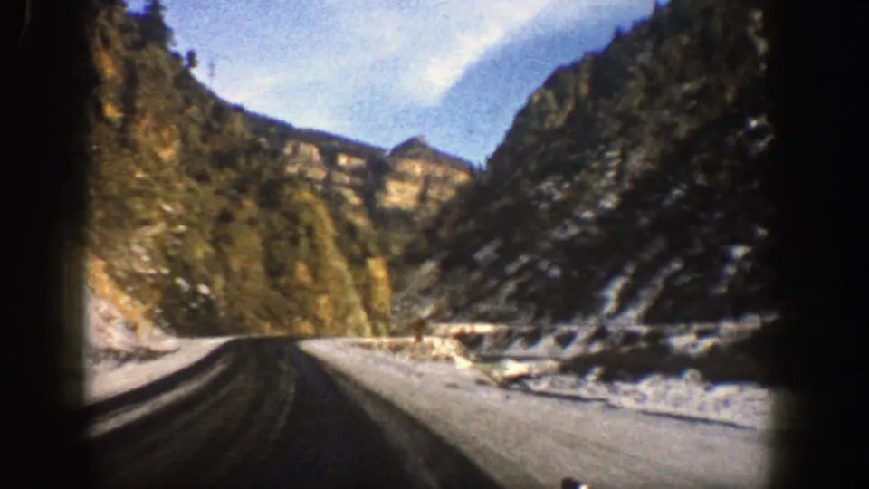 A 1961 car-view shot in Aspen, Colorado shows a paved mountain road bending between steep canyon walls with snow along the roadside.